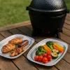 Twee aluminium serveerschalen met gegrilde kip, worst en groenten op een houten tafel naast een Kamado Ho-cooker.