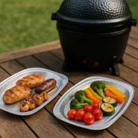 Twee aluminium serveerschalen met gegrilde kip, worst en groenten op een houten tafel naast een Kamado Ho-cooker.