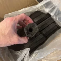 Close-up van een House of Charcoal hexagon briket – geperst hardhout met luchtkanaal, vastgehouden boven een doos vol briketten